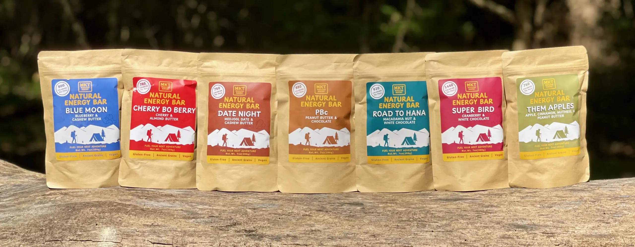 Outdoor back drop, with seven flavors of natural energy bars, standing in line on a log.  Blueberry and Cashew Butter, Cherry and Almond Butter, Medjool Date and Cashew Butter, Peanut-Butter and Chocolate, Macadamia Nut and White Chocolate, Cranberry and White Chocolate, and Apple, Cinnamon, Nutmeg, and Peanut Butter.